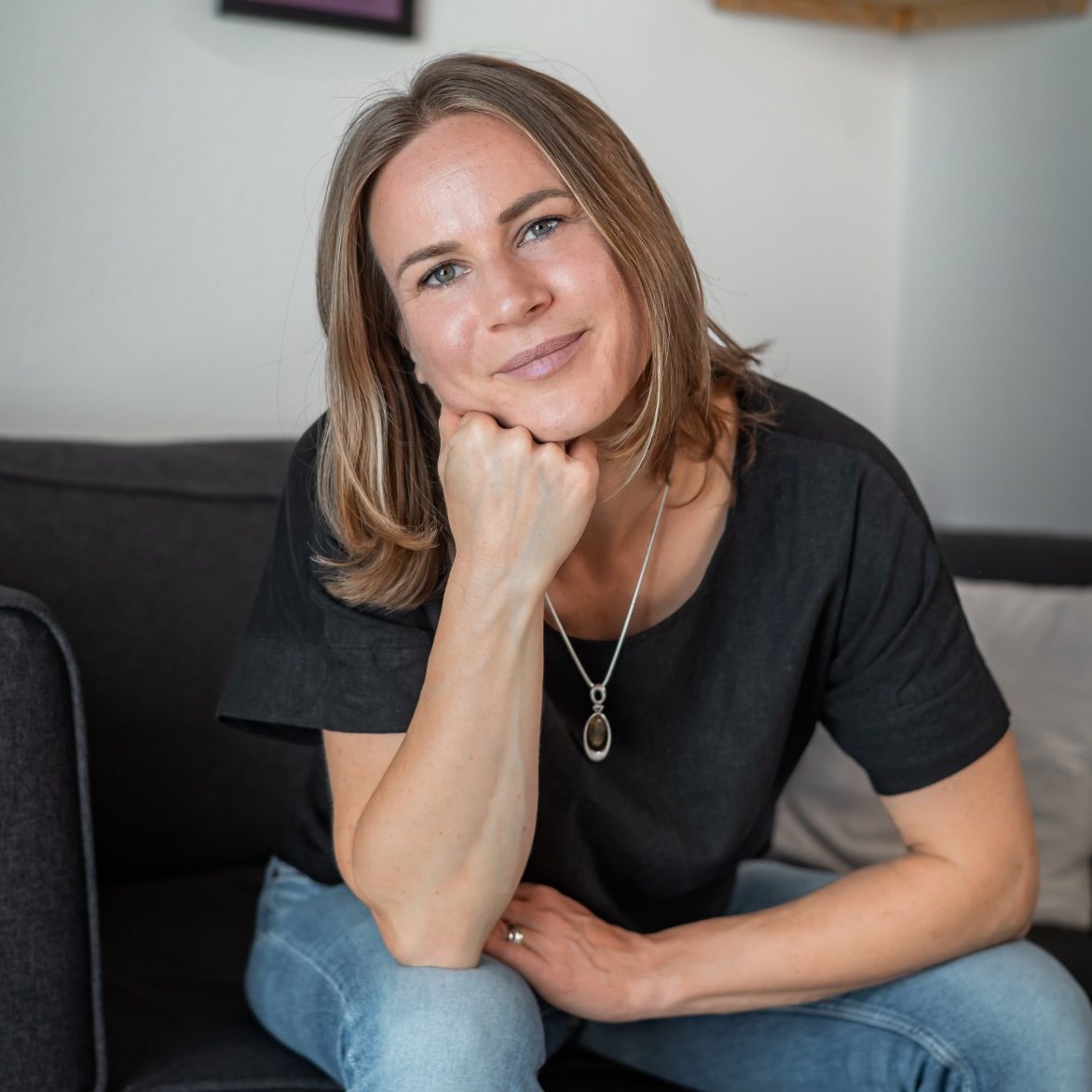 Portrait Jasmin Kircher. Kopf in Hand gestützt, lächelnd, sitzt auf Couch