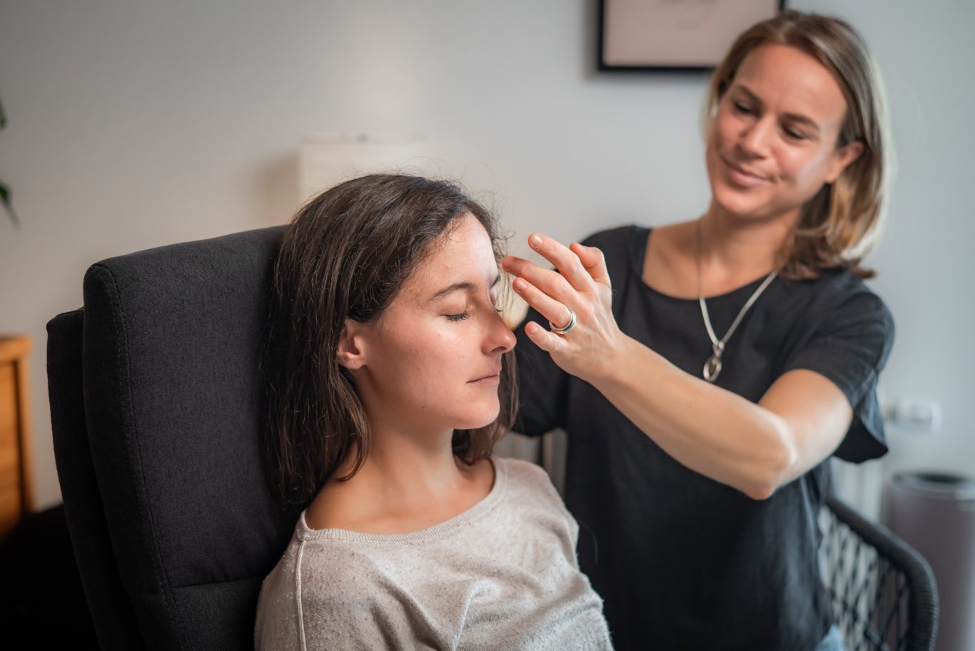 Junge Frau (Klientin) sitzt mit geschlossenen Augen. Hypnotiseurin (Jasmin Kircher) berührt sie an der Stirn.