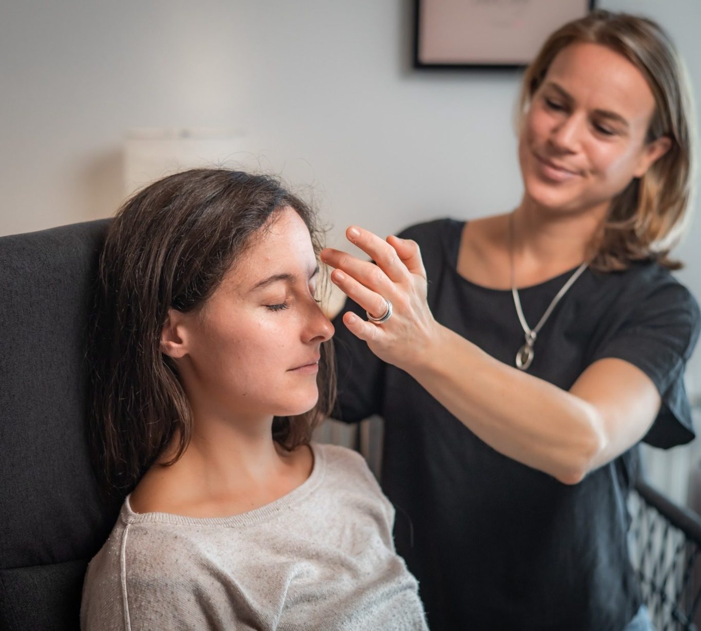 Junge Frau (Klientin) sitzt mit geschlossenen Augen. Hypnotiseurin (Jasmin Kircher) berührt sie an der Stirn.
